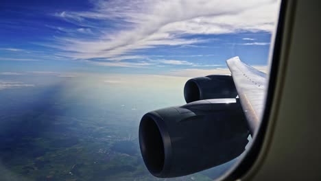 looking through the window of a passenger airliner with the engines and wing in the foreground against a blue sky with clouds.