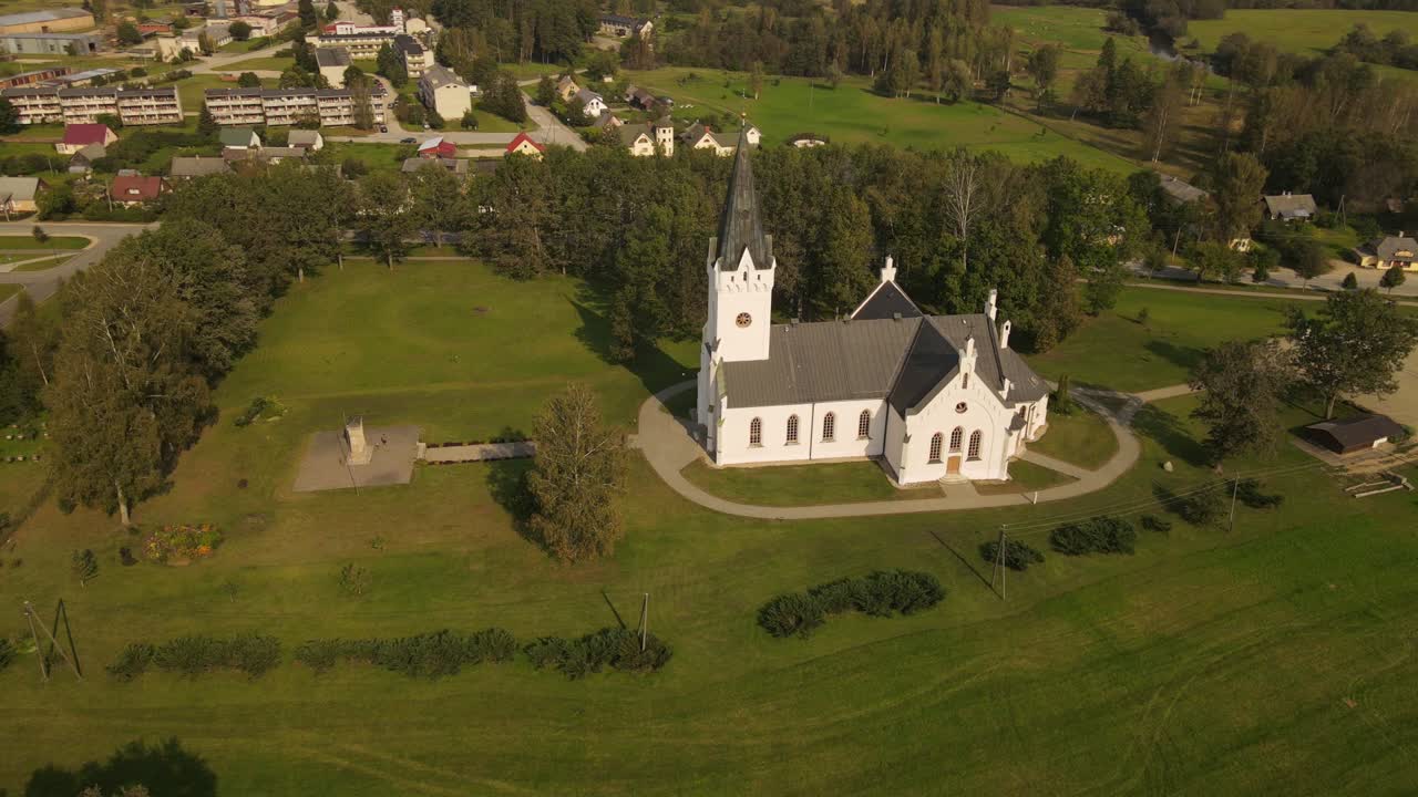 Drone view of St. Thomas Evangelical Lutheran Church in Jaunpiebalga, captured on a bright day. The church stands out among the town's buildings, surrounded by greenery and streets.