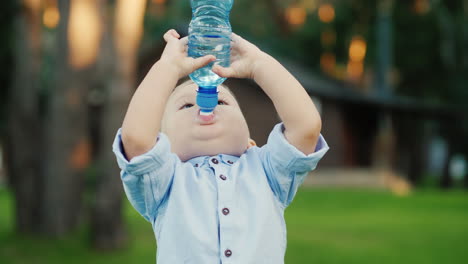 un niño divertido de 1 año está bebiendo agua de una botella que se encuentra en un césped verde y se tambalea con frialdad video divertido