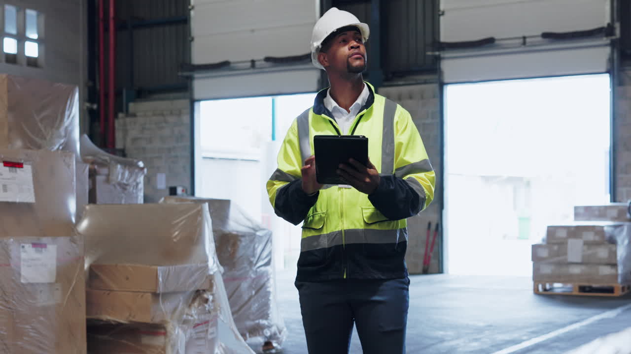 un trabajador de almacén en un chaleco de seguridad y casco utiliza una tableta para inspeccionar cajas e inventario
