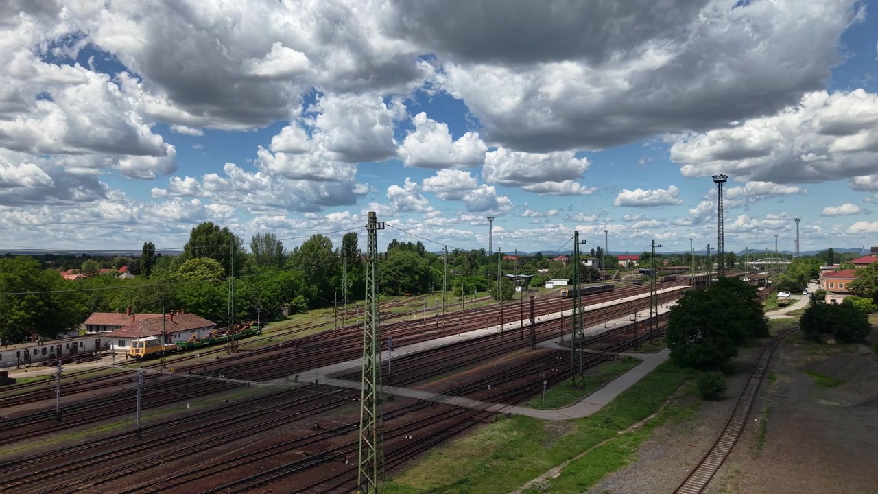 En vidvinkeludsigt over en kompleks rangerbanegård i Ungarn med mange spor under en dramatisk overskyet himmel.