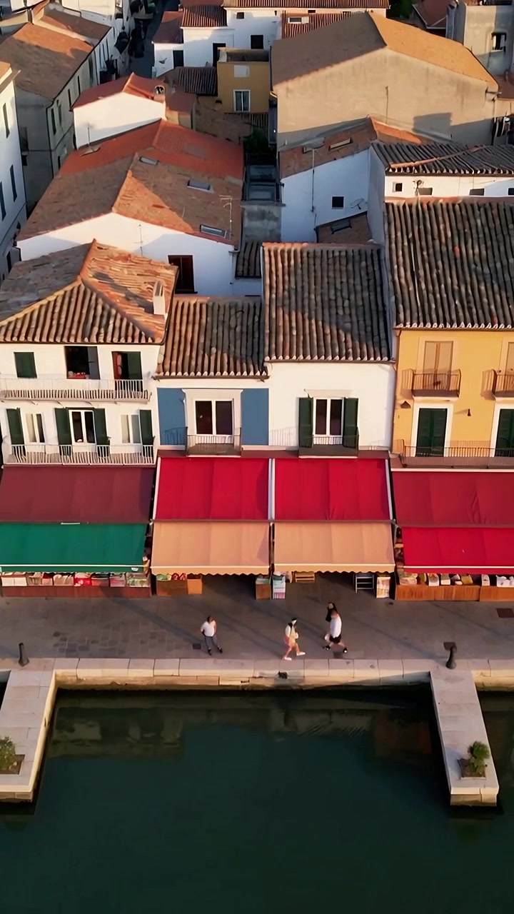 Video verticale: Telecamera scorrevole che rivela una banchina con tende da sole rosse e color cuoio, pedoni che camminano lungo il canale