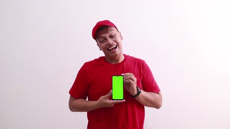 happy young asian courier in red standing while showing green screen blank mobile phone display