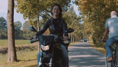 front view of beautiful smiling european female motor biker in leather jacket driving on road passing cyclists with autumn leaf color trees in forest on sunny day