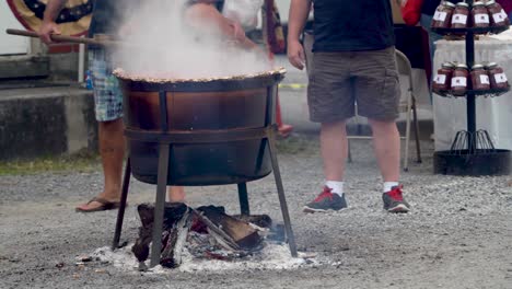 Two-men-standing-as-one-of-them-stirs-steaming-apple-butter-in-a-large-wood-fired-cauldron-outside