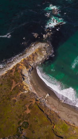 Flying over the rocky shore and moving to the azure waters of ocean. Little white waves splashing by the mountainous coastline. Top view. Vertical video