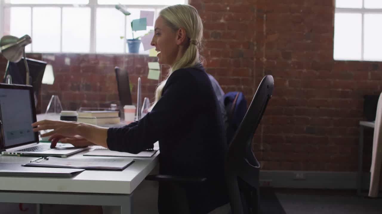 Hvid forretningskvinde sidder ved skrivebord, bruger computer og rækker et dokument til en kvindelig kollega.