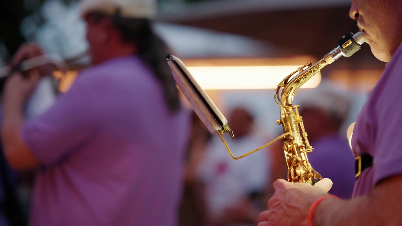 Musicista che suona un sassofono dorato, leggendo uno spartito da un leggio con altri artisti in camicia viola in una banda musicale