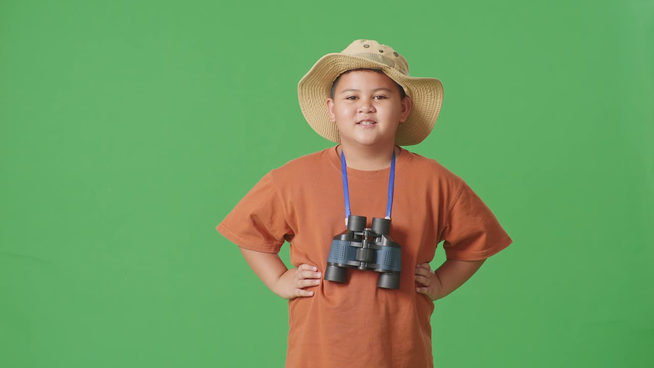 niño turista asiático con un sombrero y binoculares sonriendo a la cámara mientras está de pie con los brazos akimbo en el fondo de la pantalla verde. niño investigador examina algo, concepto de aventura de turismo de viaje