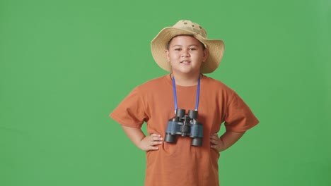 asian tourist boy with a hat and binoculars smiling to camera while standing with arms akimbo on the green screen background. boy researcher examines something, travel tourism adventure concept