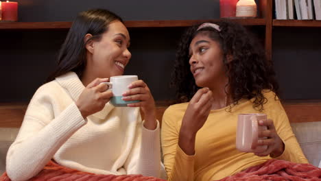 two multiracial female friends enjoying cozy conversation with mugs at home, sharing laughter
