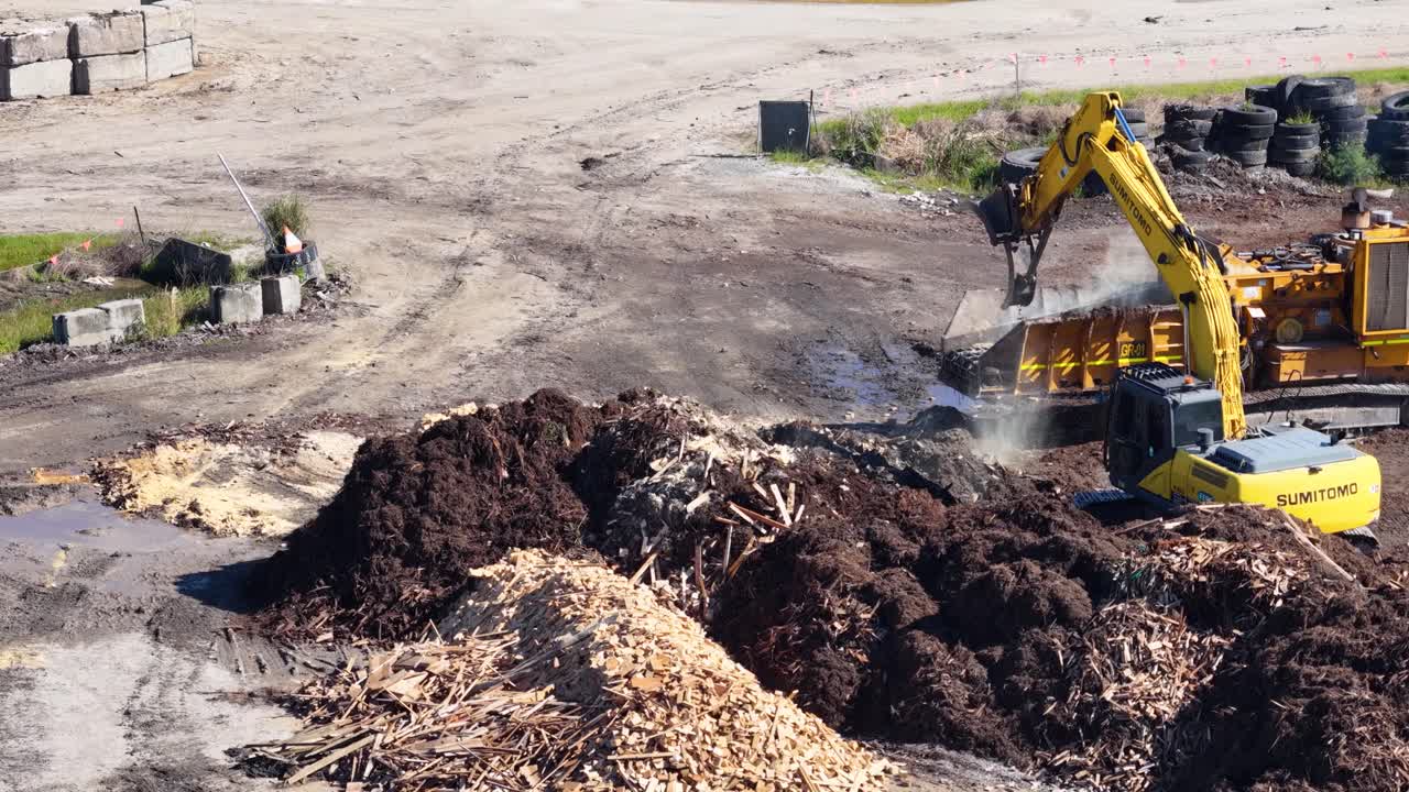 Drone captures an excavator sorting mulch and soil in an industrial yard. Bright daylight highlights the machinery and landscape
