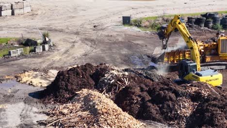 drone captures an excavator sorting mulch and soil in an industrial yard. bright daylight highlights the machinery and landscape