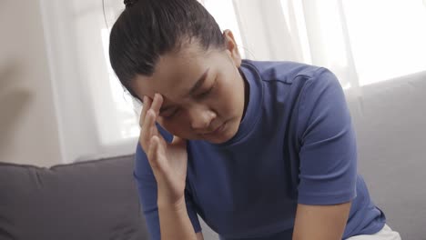 asian woman wearing a blue shirt sitting on a sofa feeling very headache