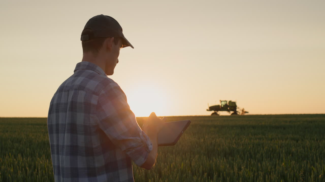 un joven agricultor exitoso está trabajando en el campo. un tractor está conduciendo en el fondo. un hombre usa una tableta - nuevas tecnologías en la agricultura
