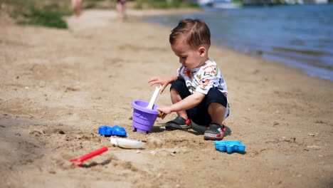 lovely toddler boy plays with toys on the river bank. kid takes some sand with shovel and puts it into bucket.