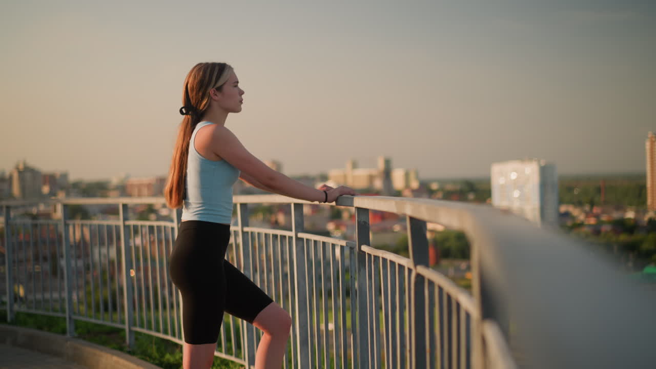 야외에 서 있는 젊은 여성, 손은 철의 레일링에 고, 따뜻한 빛 아래 멀리서 사려 깊게 쳐다보고, 배경의 도시 풍경은 효과를 위해 흐릿합니다.