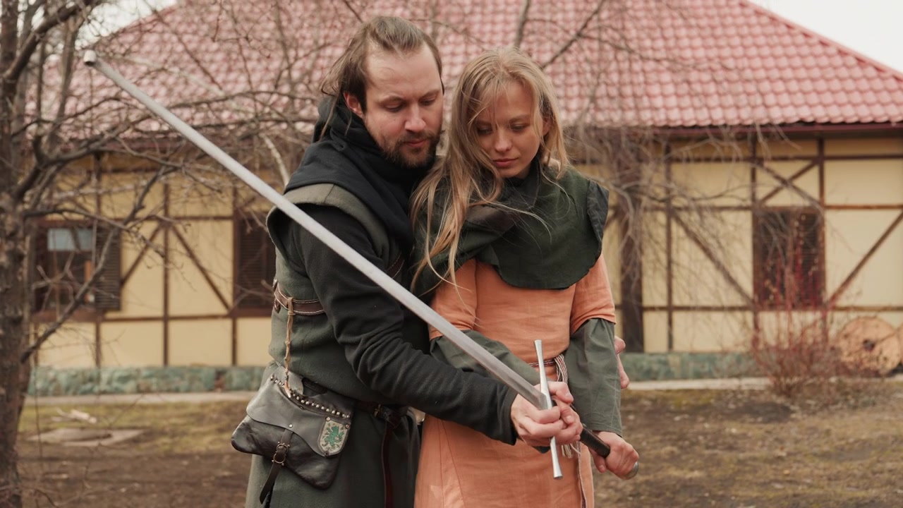Mannlige og kvinnelige reenactere står tett sammen på en gårdsplass mens mannen veileder kvinnens hender langs et langt sverd, viser kontrollert bevegelse og fokuserte uttrykk under historisk trening