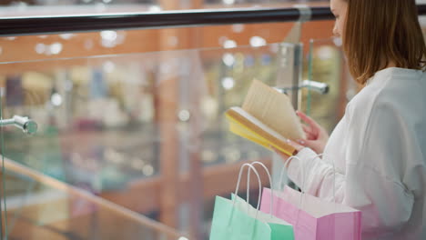 woman flipping through a yellow book while standing near railing in shopping mall, colorful shopping bags are placed on the floor, and the background features blurred glass reflections