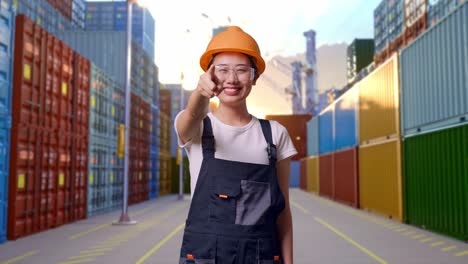 trabajadora asiática con gafas de protección y casco de seguridad sonriendo y tocando su pecho luego señalándote mientras está de pie en el almacén del patio de contenedores