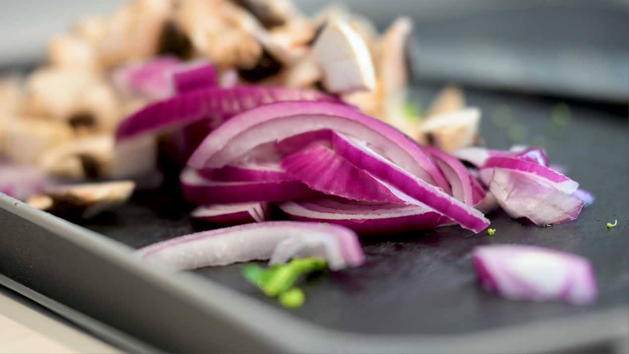 A close-up shot with a narrow focus on freshly sliced red onions on a black cutting board. Chopped mushrooms are softly blurred in the background, ready for cooking