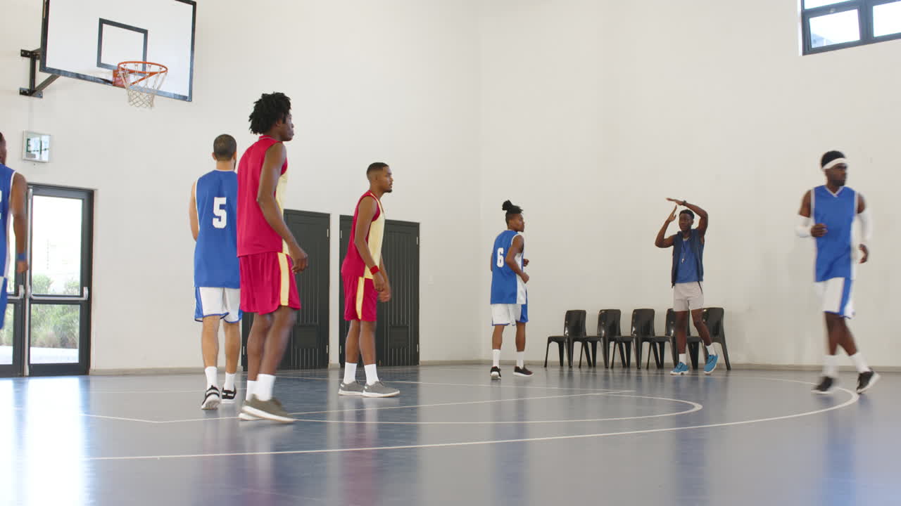 Multiracial male basketball players playing basketball, coach pausing game and team huddling in gym