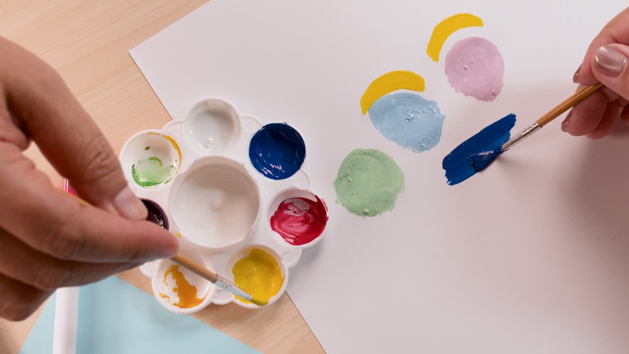 Hands paint colorful shapes on paper using brushes, vibrant palette, overhead view, natural lighting