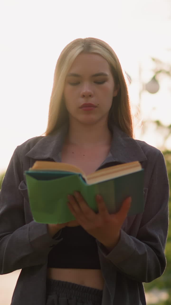 dama con ropa gris caminando por un campo de hierba leyendo un libro mientras la luz del sol crea destellos brillantes a su alrededor, con una vista borrosa de árboles y personas en el fondo