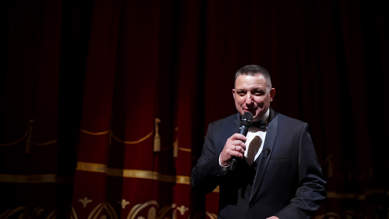 Presenter with microphone announces performance before auditorium from stage. Theater curtains background.
