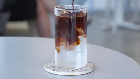 concepto de publicidad de restaurante de cafetería cinematográfica, solución diaria de cafeína, un vaso de helado largo negro con cubitos de hielo, agregando agua fría y mezcla de café expreso de la casa y revolviendo bien con una pajita de vidrio