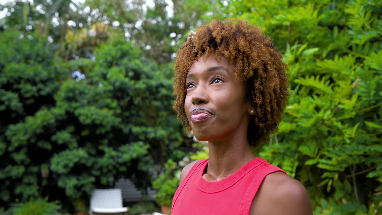 Afro-Amerikaanse vrouw in rode top in de tuin verlegt haar blik en glimlacht, reagerend op een stem buiten beeld