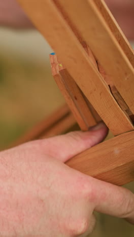 un primer plano de las manos de un pintor ajustando un trípode de madera en un campo cubierto de hierba. el área circundante está borrosa, y la pierna de una mujer es visible en el fondo, añadiendo profundidad a la escena