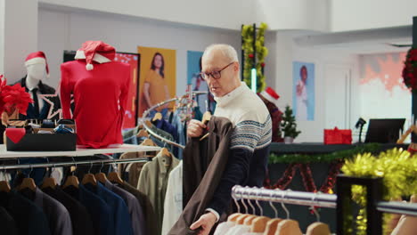 dolly out shot of elderly client in xmas decorated shopping mall store trying to determine if stylish blazer is fitting him. senior shopper in fashion boutique during christmas sales