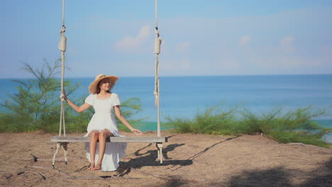 woman in long sundress and hat on swing with ocean background