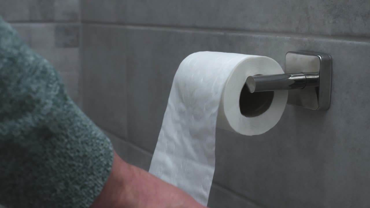 A close-up of a hand pulling a sheet from a toilet paper roll mounted on a metallic holder in a tiled bathroom. The scene highlights hygiene and daily life essentials.