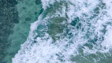 Top-Down-Slow-Motion-Drone-View-of-turquoise-clear-water-and-crashing-waves-over-shallow-coral-reef-in-Uluwatu-Bali-Indonesia