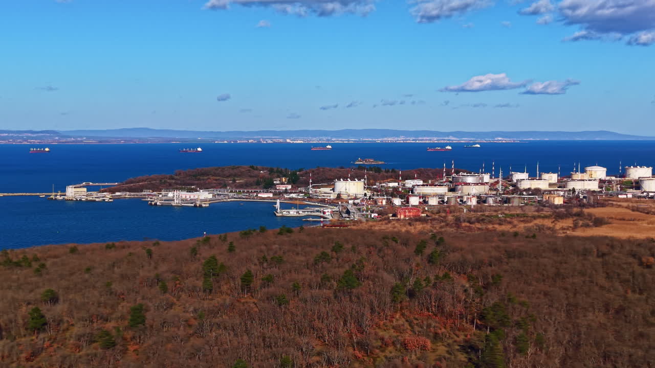 Utsikt over industriområde med tanker og skip nær kystlinjen observert på avstand, som viser land- og vannfunksjoner under en klar, lys himmel.