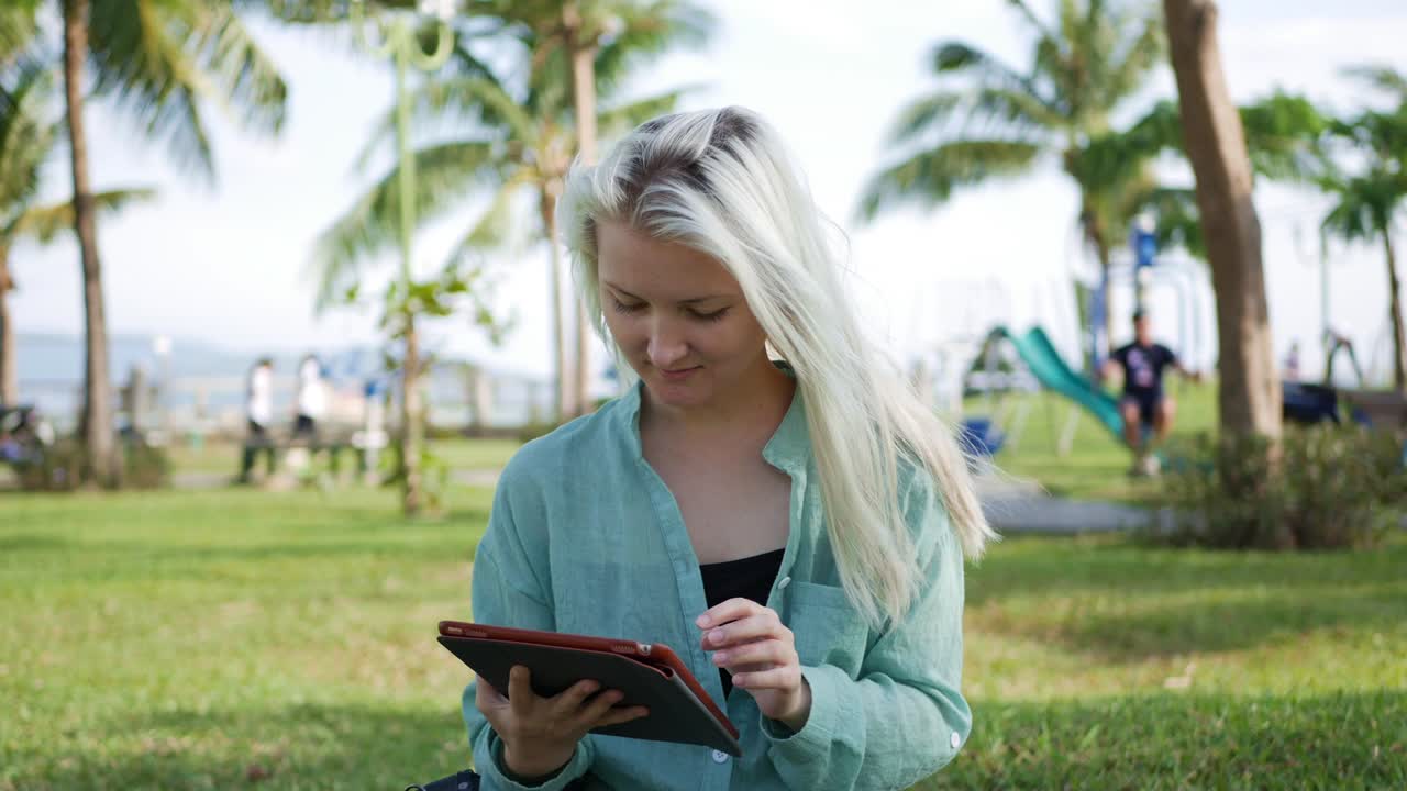Beautiful slim woman with long blonde hair in green shirt sits on the ground and using smartphone over background the park. Girl on the square touching screen and smile