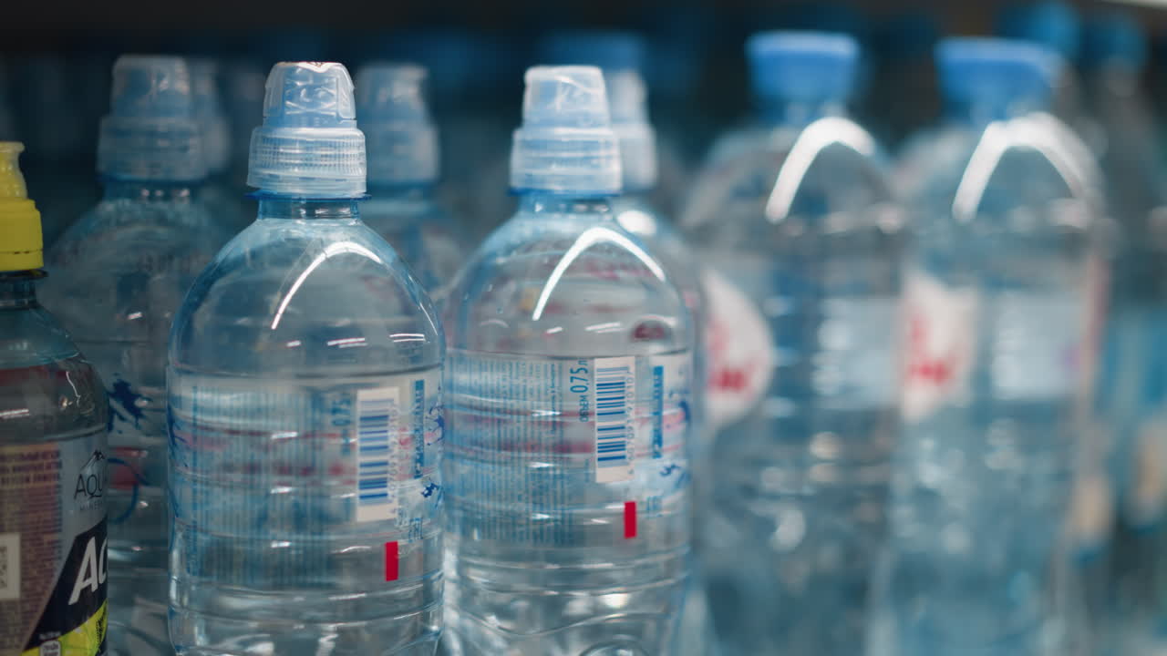 mano con guantes con cuentas tomando una botella de agua de un estante. disparado con una cámara de mano, haciendo hincapié en el acto de seleccionar una botela de agua entre muchos en un estante de la tienda