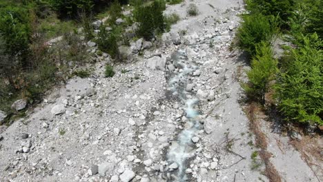 Drohnenansicht-In-Albanien-In-Den-Alpen,-Die-über-Einen-Kristallklaren-Fluss-Mit-Felsigem-Boden-Und-Grünem-Wald-An-Den-Seiten-Im-Theth-Fliegt