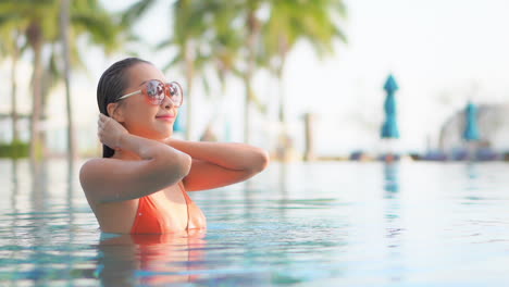 Junges-Asiatisches-Weibliches-Model-Im-Orangefarbenen-Bikini,-Das-Nasse-Haare-Im-Swimmingpool-Bei-Exotischem-Sonnenuntergang-In-Zeitlupe-Aus-Nächster-Nähe-Glättet