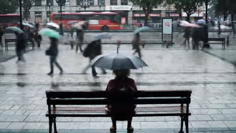 in a bustling city during a heavy rainstorm, numerous individuals navigate the streets under umbrellas while one person remains seated on a bench, seemingly lost in thought.