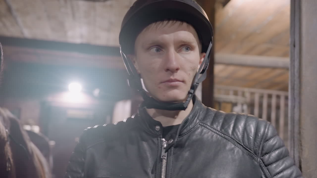 White man in stable checking helmet strap and buckles, closeup of focused face and gloved hands securing chin area, leather jacket and nearby horse visible, serious preride inspection and preparation