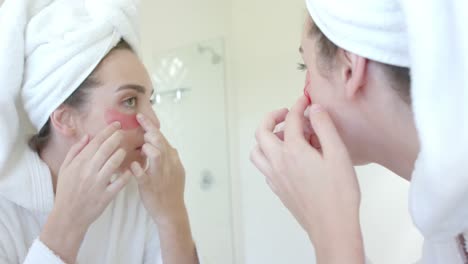 caucasian woman with towel on head applying eye patches in bathroom in slow motion