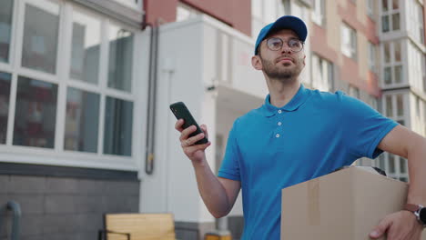 Caucasian-young-pretty-man-delivery-worker-in-blue-cap-walking-the-street-and-carrying-carton-box-while-using-smartphone-looking-for-route.-Male-courier-with-parcel-tapping-and-texting-on-phone.