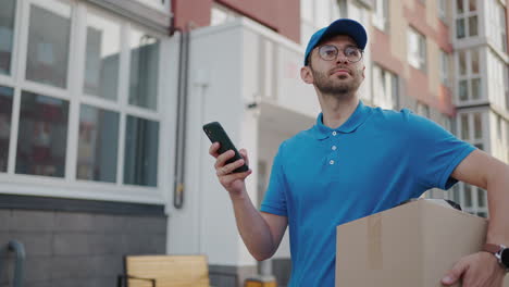 Kaukasischer-Junger,-Hübscher-Lieferarbeiter-Mit-Blauer-Mütze,-Der-Auf-Der-Straße-Läuft-Und-Einen-Karton-Trägt,-Während-Er-Sein-Smartphone-Benutzt,-Um-Nach-Der-Route-Zu-Suchen.-Männlicher-Kurier-Mit-Paketabhörung-Und-SMS-Am-Telefon.