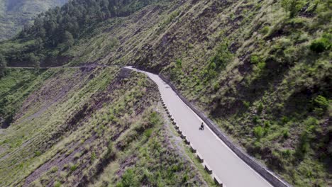 Toma-Aérea-De-Un-Hombre-En-Bicicleta-Con-Equipaje-Que-Viaja-Por-Una-Carretera-Escénica-Vacía-En-Una-Región-Montañosa-Fría-O-Pasa-Por-Exuberantes-Granjas-Verdes-Y-Un-Valle