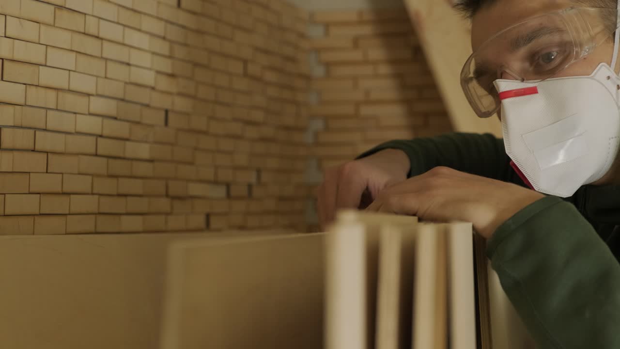 Un falegname professionista con mascherina e occhiali controlla legno grezzo, un'asse di legno e un pannello di compensato.