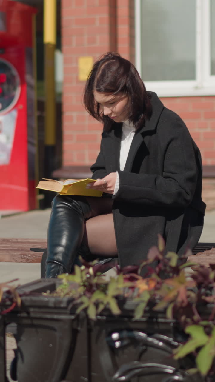 investigadora sentada al aire libre con las piernas cruzadas, centrada en el libro en sus manos. en el fondo, una máquina hace sonar mientras las flores en una olla se balancean, en un día soleado