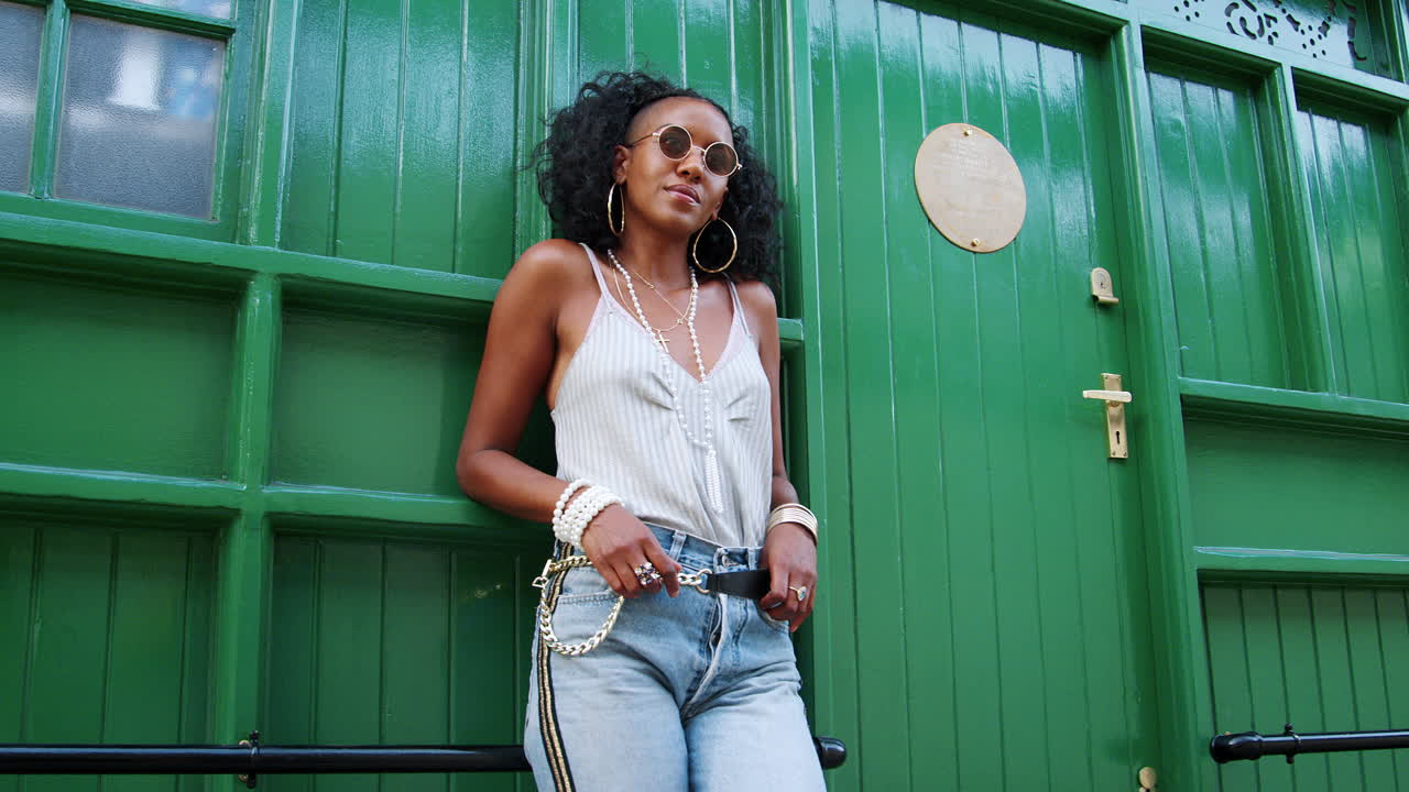 Fashionable young black woman wearing sunglasses leaning by green door outdoors looking at camera, low angle